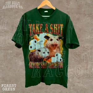 A green t-shirt with a graphic design featuring two opossums and toilet paper rolls, along with the text "TAKE A SHIT BEFORE SHIT TAKES YOU".