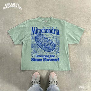 A person's feet wearing white and black sneakers are standing on a concrete floor, with a green t-shirt featuring a graphic design and text printed on it.