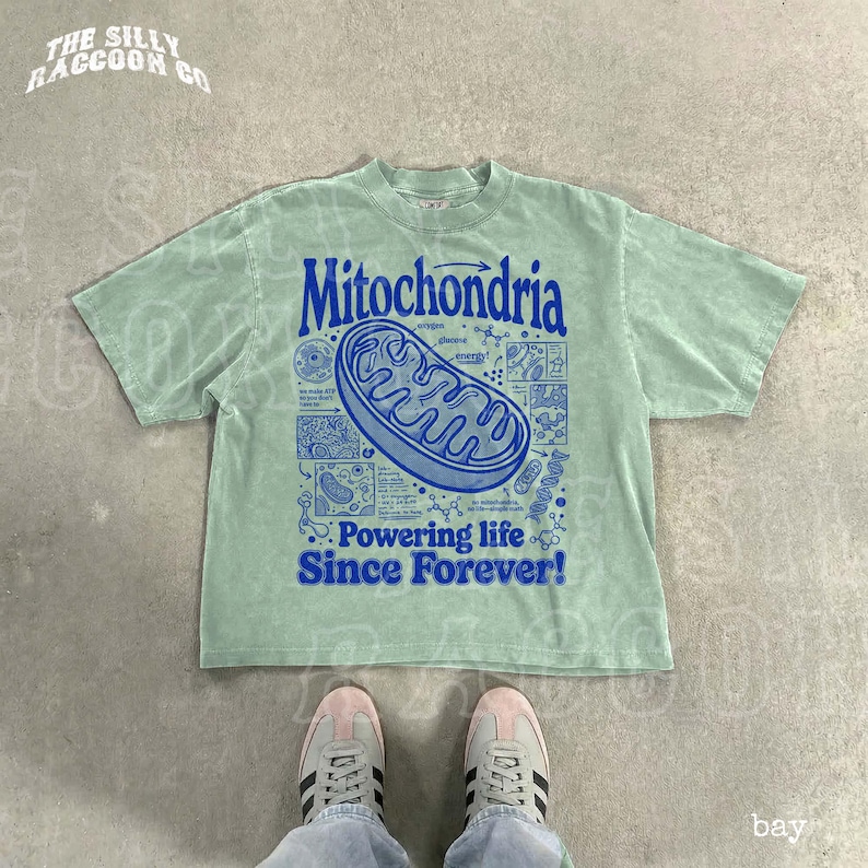 A person's feet wearing white and black sneakers are standing on a concrete floor, with a green t-shirt featuring a graphic design and text printed on it.