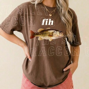 A woman wearing a brown t-shirt with a fish graphic and red and white shorts, smiling at the camera.