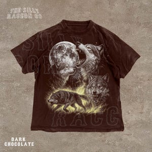 a t-shirt with a graphic design depicting three wolves, one of which is howling at the moon. The wolves are shown in a dark brown color, and the moon is depicted as a large, glowing orb. The t-shirt is displayed against a beige background.