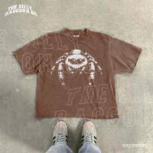 A brown t-shirt with a white spider graphic printed on it, laid out on a concrete floor. A pair of white sneakers is visible underneath the t-shirt.