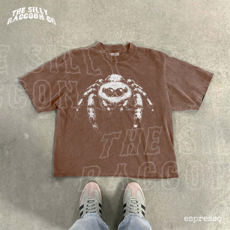 A brown t-shirt with a white spider graphic printed on it, laid out on a concrete floor. A pair of white sneakers is visible underneath the t-shirt.