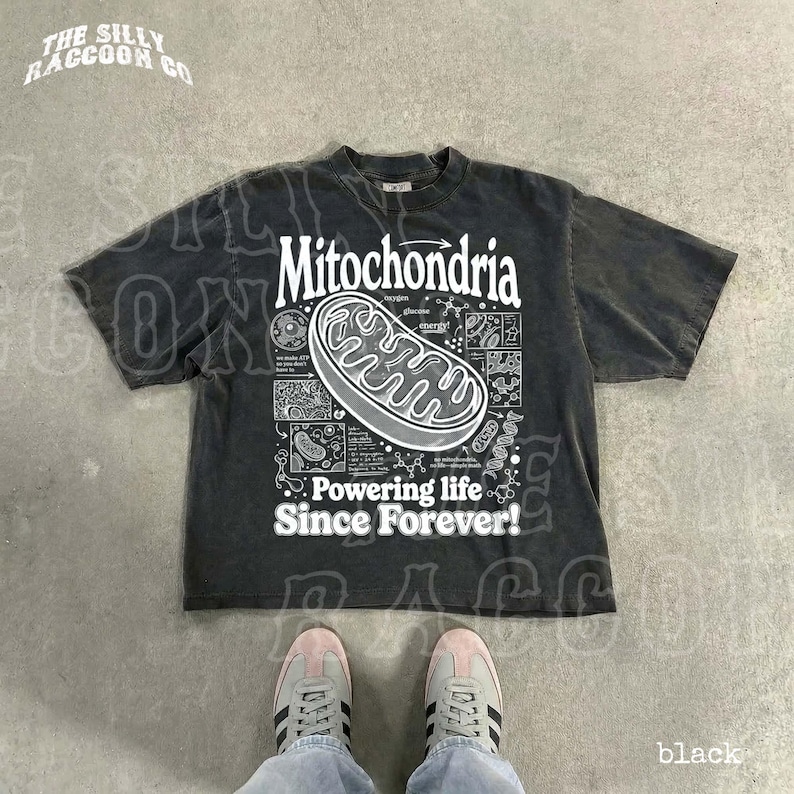 a black t-shirt with a graphic design depicting Mitochondria, a cellular organelle, and the text "Powering life since forever!" printed on it. The t-shirt is laid out on a concrete floor, and a pair of feet is visible in the bottom right corner of the image.