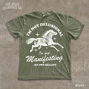 A green t-shirt with a graphic design depicting a horse and the text "I'm not delusional, I'm just Manifesting MY OWN REALITY" printed on it.