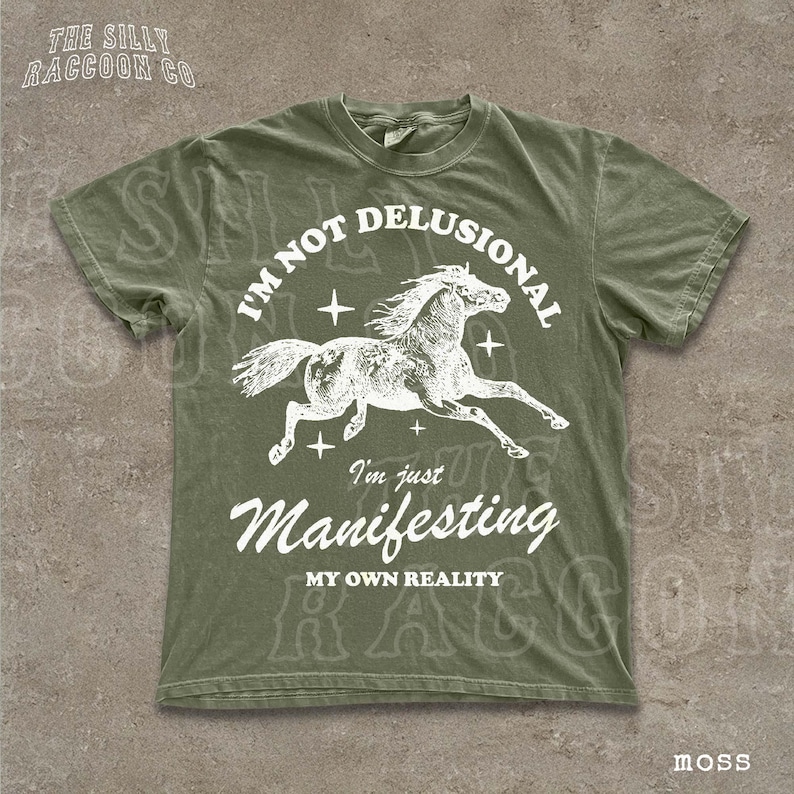 A green t-shirt with a graphic design depicting a horse and the text "I'm not delusional, I'm just Manifesting MY OWN REALITY" printed on it.