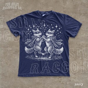 A navy blue t-shirt with a graphic design featuring two foxes dancing under a starry sky.
