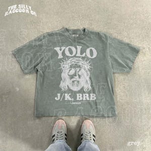 A grey t-shirt with a graphic design and text printed on it, laid out on a concrete floor.