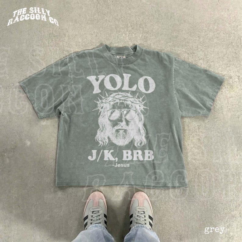 A grey t-shirt with a graphic design and text printed on it, laid out on a concrete floor.