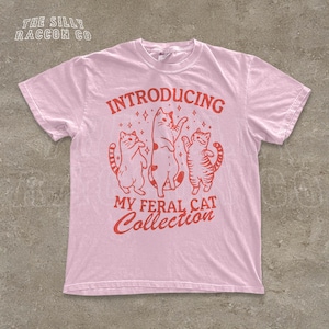 A pink t-shirt with a graphic design featuring three cats and the text "INTRODUCING MY FERAL CAT COLLECTION" printed on it.