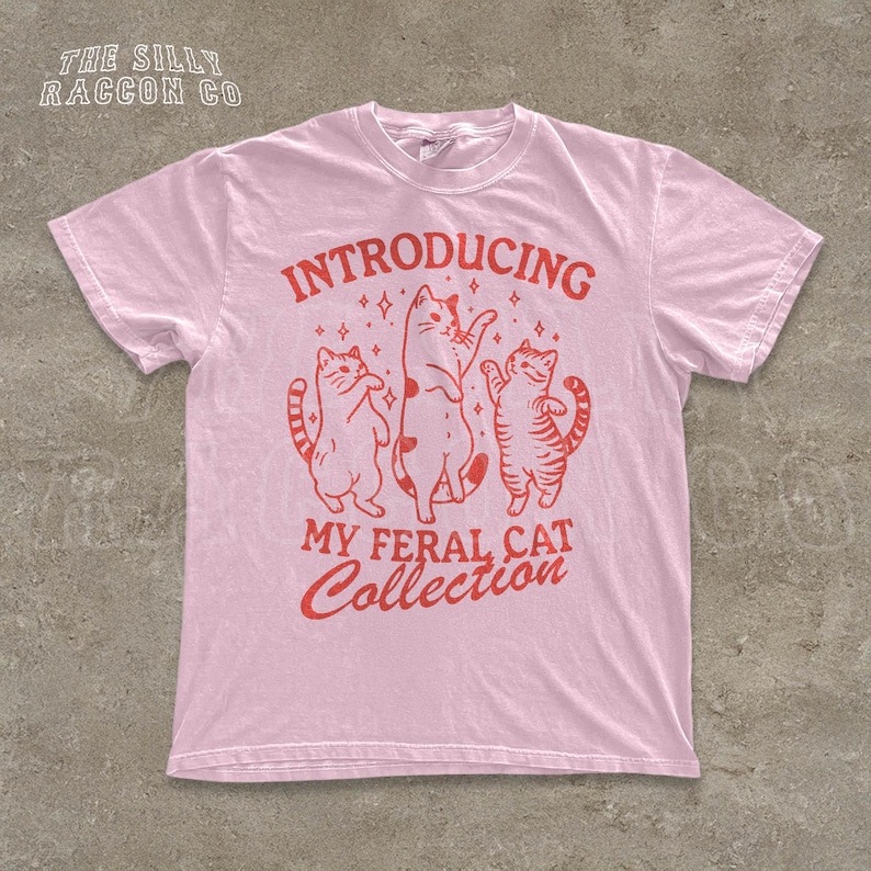 A pink t-shirt with a graphic design featuring three cats and the text "INTRODUCING MY FERAL CAT COLLECTION" printed on it.