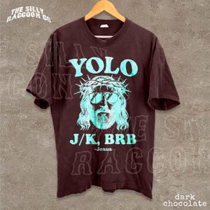 A dark brown t-shirt with a graphic design featuring a stylized depiction of Jesus wearing sunglasses and a crown, along with the text "YOLO J/K. BRB Jesus" printed on it.