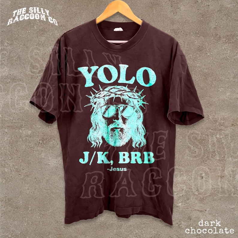 A dark brown t-shirt with a graphic design featuring a stylized depiction of Jesus wearing sunglasses and a crown, along with the text "YOLO J/K. BRB Jesus" printed on it.