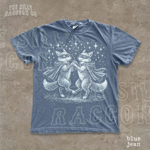 A blue t-shirt with a graphic design featuring two raccoons dancing under a starry sky.