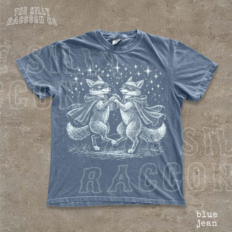 A blue t-shirt with a graphic design featuring two raccoons dancing under a starry sky.