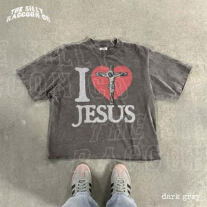 A gray t-shirt with a graphic design of a cross and a heart, along with the text "I ♥ JESUS" printed on it. The t-shirt is laid out on a concrete floor, and a pair of sneakers is visible underneath it.