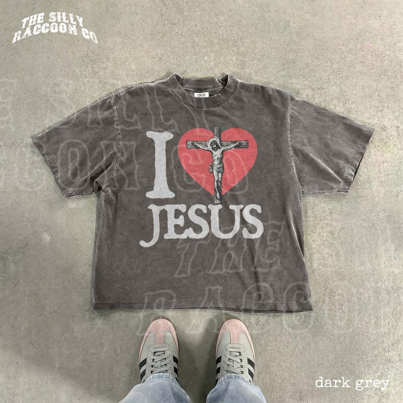 A gray t-shirt with a graphic design of a cross and a heart, along with the text "I ♥ JESUS" printed on it. The t-shirt is laid out on a concrete floor, and a pair of sneakers is visible underneath it.