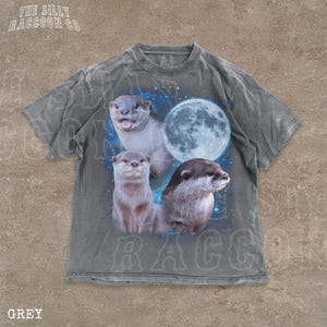 A grey t-shirt with a graphic design featuring three otters against a backdrop of a full moon and stars.