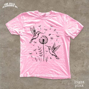A pink t-shirt with a graphic design featuring two hummingbirds, a dandelion, and other floral elements.
