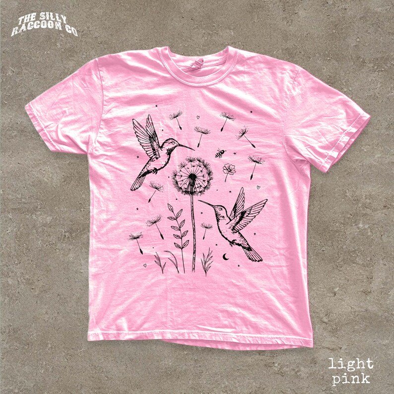 A pink t-shirt with a graphic design featuring two hummingbirds, a dandelion, and other floral elements.