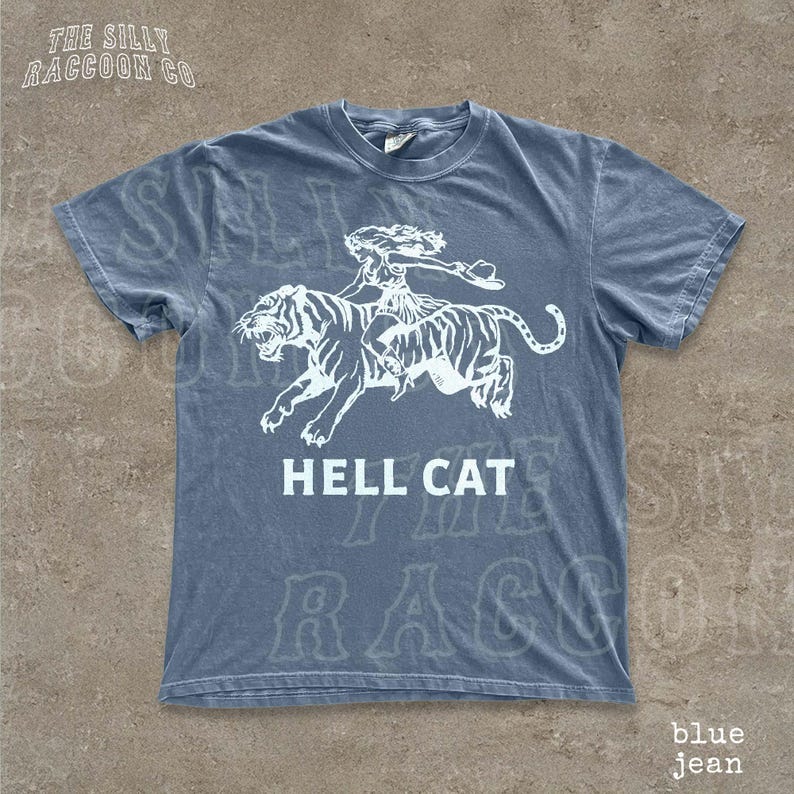 A blue t-shirt with a graphic design depicting a tiger and a person riding on its back, along with the text "HELL CAT" printed on it.
