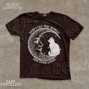 a black t-shirt with a graphic design depicting a cat sitting on a crescent moon and a human face. The text on the t-shirt reads "Don't let the world dim your light".
