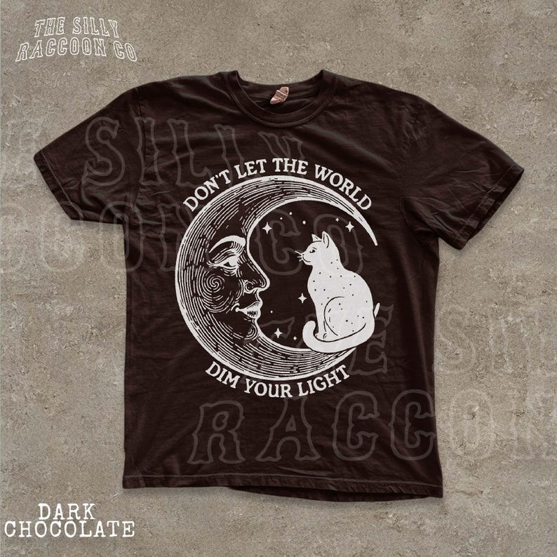 a black t-shirt with a graphic design depicting a cat sitting on a crescent moon and a human face. The text on the t-shirt reads "Don't let the world dim your light".