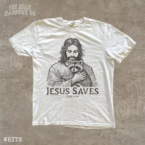 A white t-shirt with a graphic design depicting a man holding a raccoon. The text below the graphic says "JESUS SAVES LUKE 19:10".