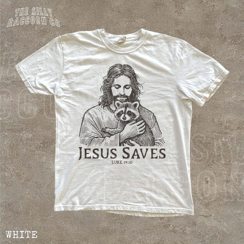 A white t-shirt with a graphic design depicting a man holding a raccoon. The text below the graphic says "JESUS SAVES LUKE 19:10".