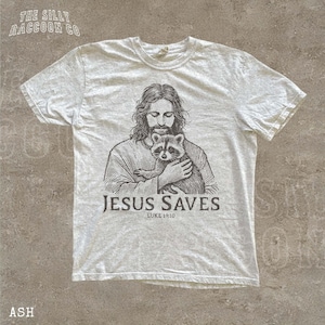 A white t-shirt with a graphic design depicting a man holding a raccoon. The text below the graphic says "JESUS SAVES" and "LIKE:10".