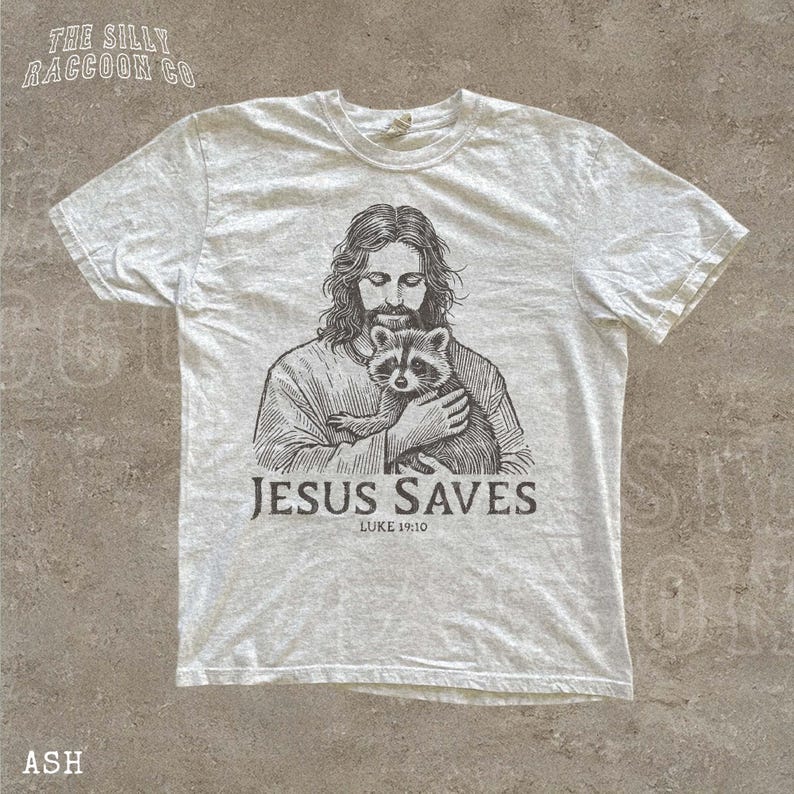 A white t-shirt with a graphic design depicting a man holding a raccoon. The text below the graphic says "JESUS SAVES" and "LIKE:10".