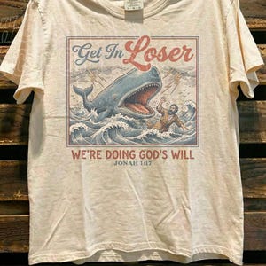 A t-shirt with a graphic design depicting a person being attacked by a large whale, with the text "Get In Loser" and "We're Doing God's Will" printed on it.