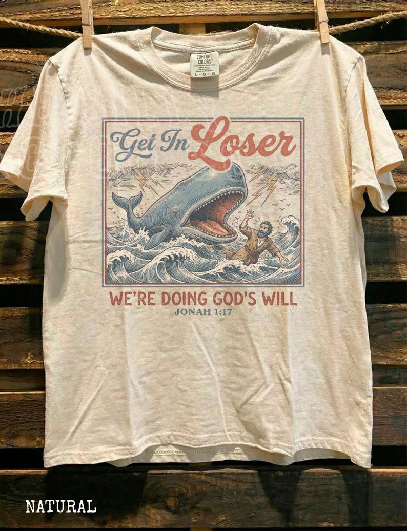 A t-shirt with a graphic design depicting a person being attacked by a large whale, with the text "Get In Loser" and "We're Doing God's Will" printed on it.