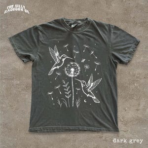 A dark grey t-shirt with a white graphic design depicting two hummingbirds hovering over a dandelion surrounded by flowers and plants.