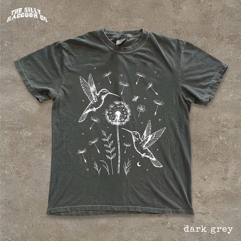 A dark grey t-shirt with a white graphic design depicting two hummingbirds hovering over a dandelion surrounded by flowers and plants.