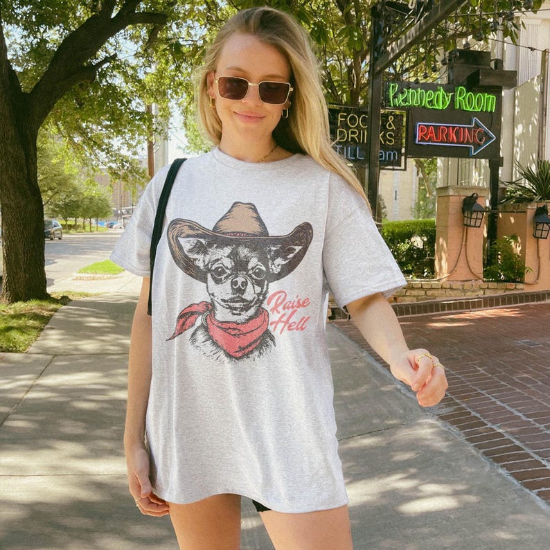 Vintage-style cowboy dog t-shirt for men and women, featuring a hand-drawn chihuahua wearing a cowboy hat and red bandana with the text "Raise Hell." Designed in a retro western aesthetic perfect for dog lovers, cowboys, and humor fans. Ideal gift for pet owners, ranch life enthusiasts, or anyone who loves funny animal graphic tees. Great for birthdays, parties, or casual country style.