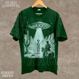 A green t-shirt with a graphic design depicting an alien spacecraft hovering over a desert landscape, with a cowboy on a horse in the foreground.