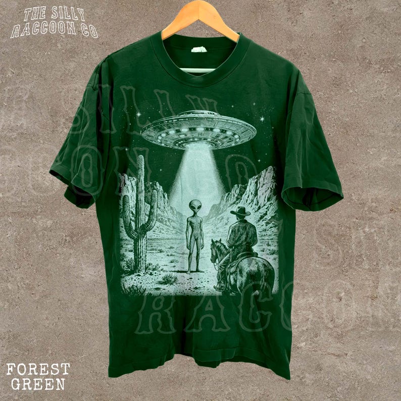 A green t-shirt with a graphic design depicting an alien spacecraft hovering over a desert landscape, with a cowboy on a horse in the foreground.