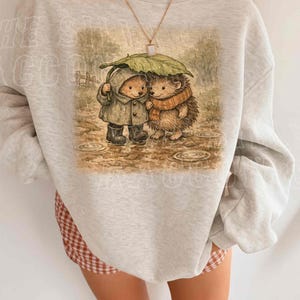 A person wearing a gray sweatshirt with a printed image of two hedgehog characters standing together under an umbrella.