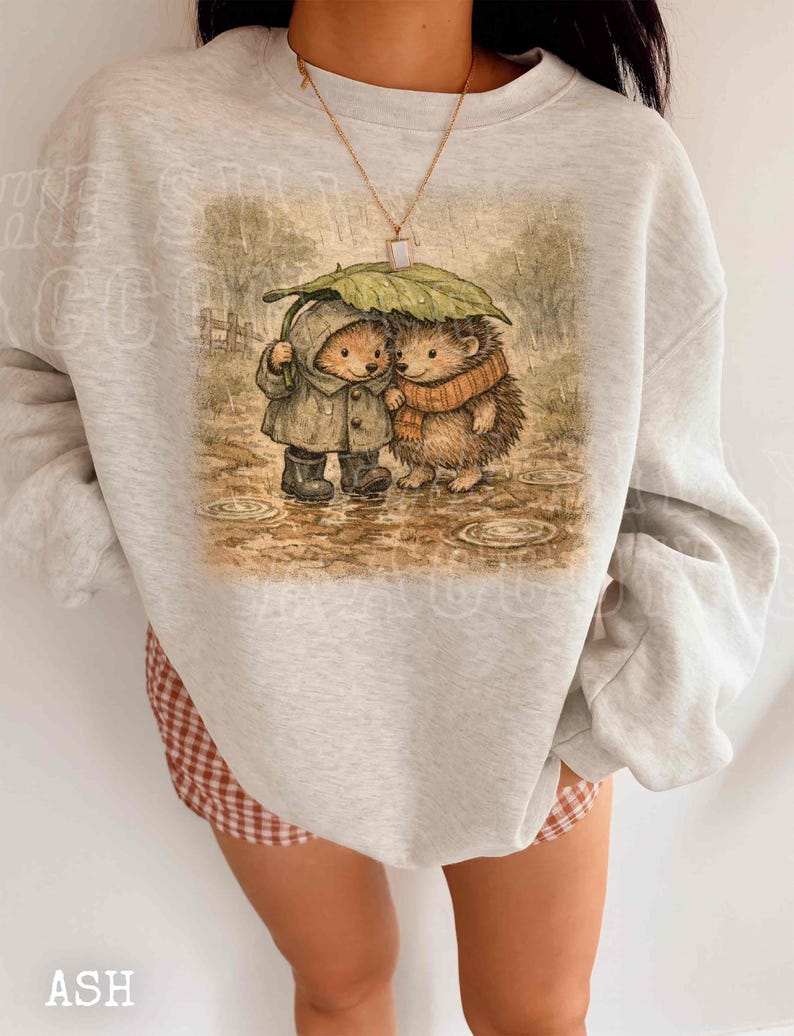 A person wearing a gray sweatshirt with a printed image of two hedgehog characters standing together under an umbrella.