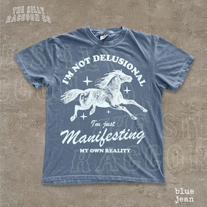 A blue t-shirt with a graphic design depicting a horse in mid-gallop, along with the text "I'm not delusional, I'm just Manifesting my own reality" printed on it.