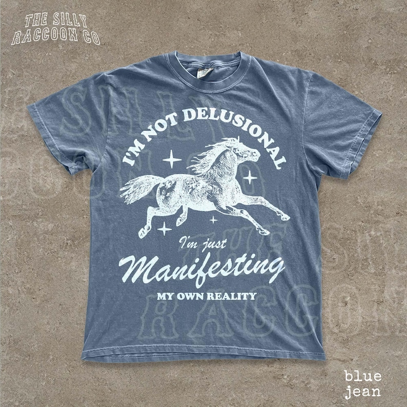 A blue t-shirt with a graphic design depicting a horse in mid-gallop, along with the text "I'm not delusional, I'm just Manifesting my own reality" printed on it.