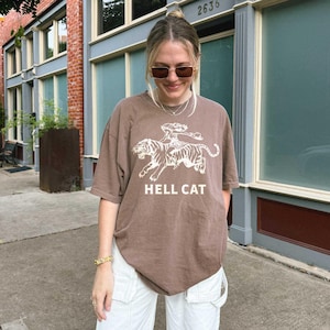 A white t-shirt featuring a striking red graphic of a woman riding a fierce tiger, with the text HELL CAT boldly printed below. The design exudes strength and wildness, making it perfect for those who love bold, adventurous styles.