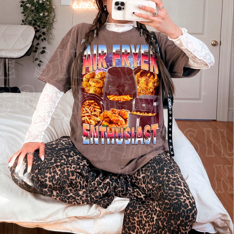 A person is sitting on a bed, wearing a t-shirt with a graphic of an air fryer and the text "AIR FRYER ENTHUSIAST" printed on it.