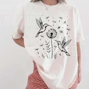 A woman wearing a white t-shirt with a graphic design of two hummingbirds and a dandelion, along with other floral and plant elements.