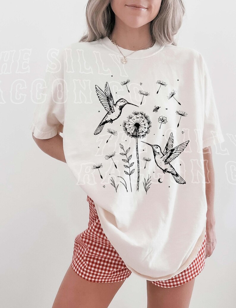 A woman wearing a white t-shirt with a graphic design of two hummingbirds and a dandelion, along with other floral and plant elements.