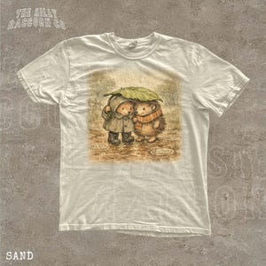 A white t-shirt with a printed illustration of two hedgehog characters standing together under an umbrella.