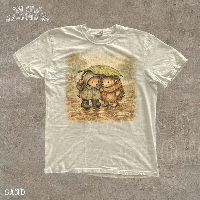 A white t-shirt with a printed illustration of two hedgehog characters standing together under an umbrella.