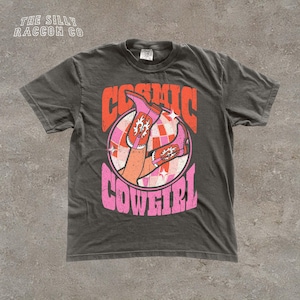 A gray t-shirt with a graphic design featuring a pair of legs and the text "COSMIC COWGIRL" in a retro style.