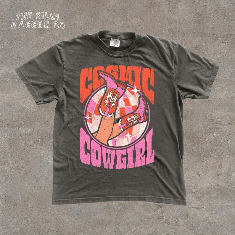 A gray t-shirt with a graphic design featuring a pair of legs and the text "COSMIC COWGIRL" in a retro style.